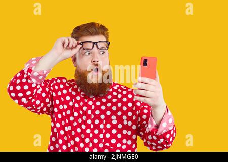 Divertente uomo chuby con l'espressione scioccata guarda lo schermo del telefono cellulare, sollevando i suoi occhiali. Foto Stock