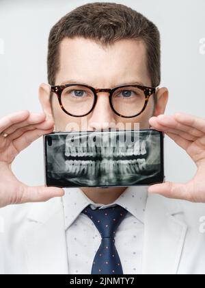 Dentista maschio coprire la bocca con uno smartphone con raggi X dei suoi denti su uno schermo di dispositivo. Foto Stock