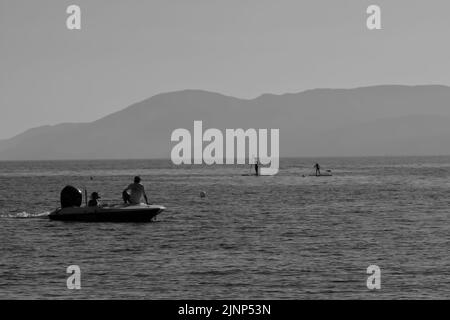 IOS, Grecia - 8 giugno 2021 : Un uomo su una piccola barca che osserva due turisti che si godono in standup paddle boarding alla spiaggia di Mylopotas in iOS Grecia in Foto Stock