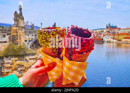 Tradizionale torta trdelnik ceca sputata, ricoperta di cioccolato e frutti di bosco secchi di fronte al fiume Moldava e pietra scolpita Ponte Carlo, Praga, CZE Foto Stock