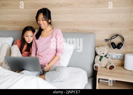 Madre e figlia asiatica che usano un computer portatile mentre si siedono a letto a casa Foto Stock