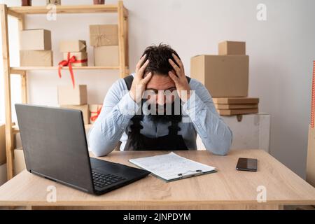 Uomo d'affari frustrato che guarda il notebook tenendo le mani a testa in frustrazione. Ritratto di stanco malato sconvolto il giovane indiano con forte emicrania Foto Stock