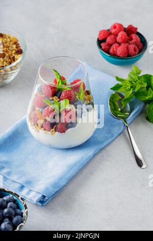 Colazione sana in un bicchiere di parfait. Yogurt con muesli e bacche su un tovagliolo blu. Lamponi freschi e mirtilli con muesli fatti in casa e y Foto Stock