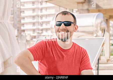 Ritratto del giovane felice milenne che sorride indossando occhiali da sole e T-shirt rossa nelle giornate estive soleggiate su sfondo luminoso Foto Stock