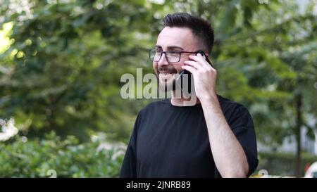 Ritratto di un giovane bell'uomo che parla al telefono con barba e occhiali. Giovane uomo sorridente in camicia nera. Sfondo natura all'aperto Foto Stock