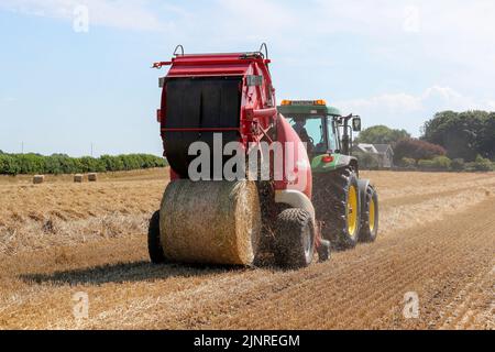 Il contadino locale guida il trattore e l'imballatrice John Dere per raccogliere e imballare il grano da tagliare per immagazzinare l'animale da lettiera e l'uso agricolo generale in inverno. N Foto Stock