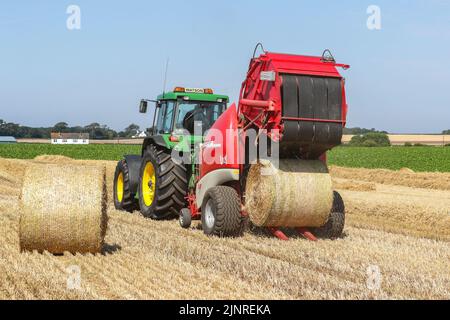 Il contadino locale guida il trattore e l'imballatrice John Dere per raccogliere e imballare il grano da tagliare per immagazzinare l'animale da lettiera e l'uso agricolo generale in inverno. N Foto Stock