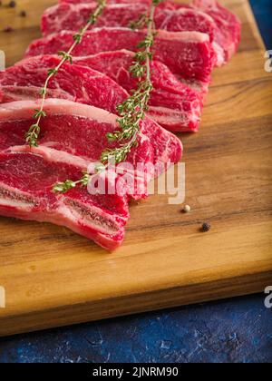 Primo piano. Bistecche di manzo crudo con fragranti rametti di rosmarino su un tagliere di legno aspettano di essere cucinate. Cibo sfondo. Ristorante, hotel, caffetteria, Foto Stock