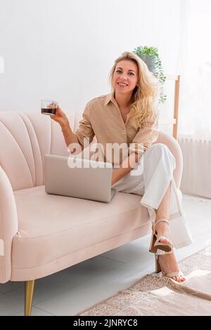 Una donna bionda, con un computer portatile grigio aperto sul divano con coppa COFee Foto Stock