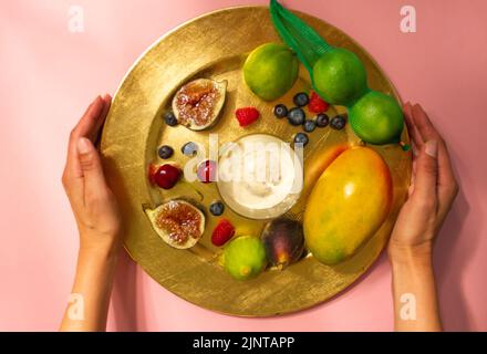Le mani della donna servono un piatto servito splendidamente, piatto di frutta tropicale fresca su un tavolo rosa vista dall'alto. Ragazza senza volto che serve sul tavolo. Fig., ma Foto Stock