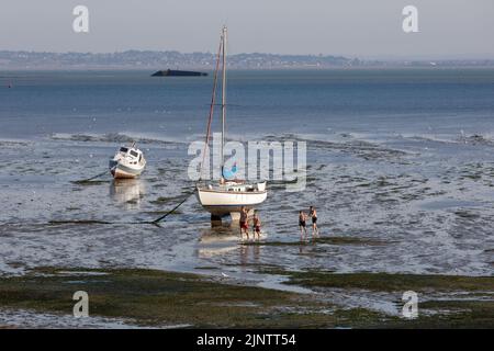 Quattro ragazzi camminano tra le barche con la bassa marea sulla costa dell'Essex, sull'estuario del Tamigi, con la costa del Kent visibile sullo sfondo Foto Stock