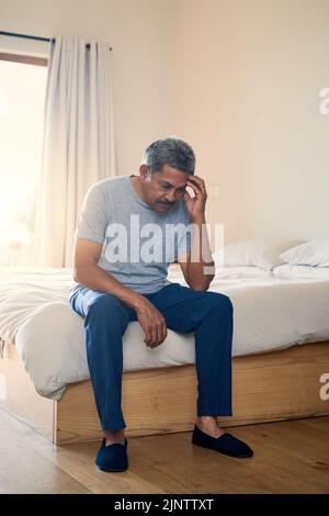 Questo mal di testa sta diventando molto irritante. Un uomo maturo stressato che tiene la testa in disagio mentre è seduto sul suo letto a casa. Foto Stock