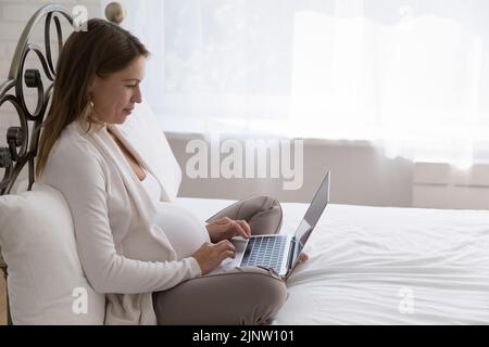 Una giovane donna incinta siede sul letto con un computer portatile Foto Stock