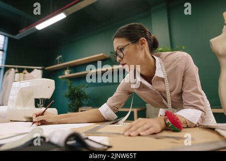 Designer di moda che prepara bozze di design su carta in studio. Donna asiatica dresssaker lavorando sul tavolo. Foto Stock