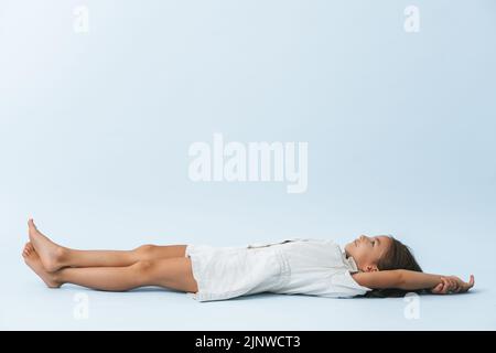 Vista laterale di una ragazza da otto anni sognante sdraiata sul pavimento, guardando verso l'alto. Su sfondo bianco bluastro. Indossa un abito corto. Foto Stock