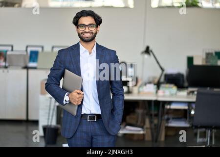 Giovane uomo d'affari indiano sorridente che indossa il vestito in piedi in ufficio, ritratto. Foto Stock