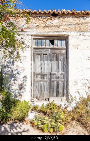 Facciata a muro di un vecchio edificio rurale, piastrelle in ceramica sul tetto, porta chiusa in legno invecchiata con vetri rotti sulla parte superiore. Casa abbandonata, piante secche e verdi fuori Foto Stock