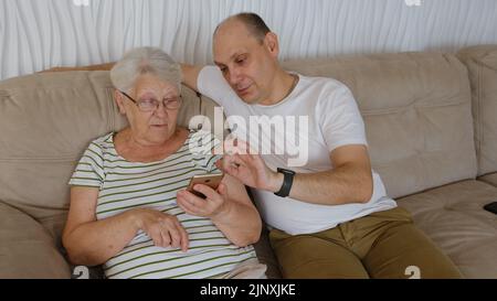 Nel soggiorno su un lato del divano seduta anziana 80s nonna con adulto 40s figlio, donna con smartphone mostra al vecchio uomo nuova app, teac Foto Stock