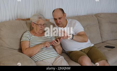 Nel soggiorno su un lato del divano seduta anziana 80s nonna con adulto 40s figlio, donna con smartphone mostra al vecchio uomo nuova app, teac Foto Stock