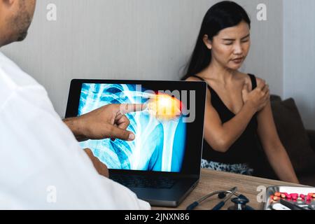 Medico che mostra una radiografia di dolore alle spalle di un notebook. Donna paziente che tiene la spalla Foto Stock