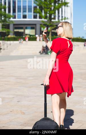 Vista posteriore di una giovane ragazza sicura in vestito rosso che cammina attraverso una città con la sua valigia e utilizzando un telefono cellulare per scattare una fotografia Foto Stock