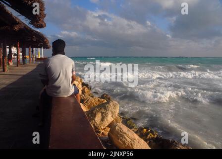 Un colpo di vista posteriore di un uomo seduto su un parapetto mentre guarda il ruvido Mar dei Caraibi, e le onde che si schiantano sulle rocce in Messico: Fuoco selettivo. Foto Stock