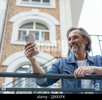 Felice uomo bearded grigio di mezza età ridendo leggendo un SMS o un divertente post sui social media utilizzando uno smartphone in piedi nella città vecchia indossando casual. Concetto di viaggio. Foto Stock