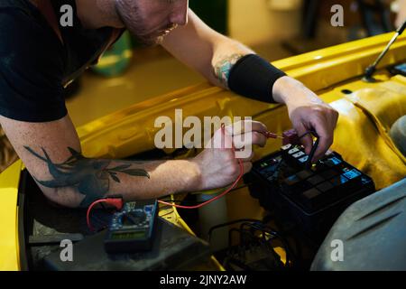 Mani di un giovane tecnico che si piega sul motore dell'auto e usa il multimetro durante il test di nuove attrezzature dopo la riparazione Foto Stock