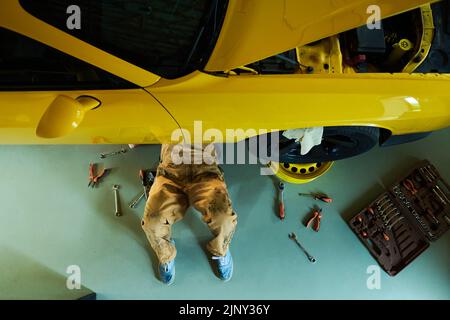 Vista dall'alto di gambe di meccanico o riparatore che fissa i dettagli di auto mentre sdraiato sul pavimento sotto auto gialla in officina Foto Stock