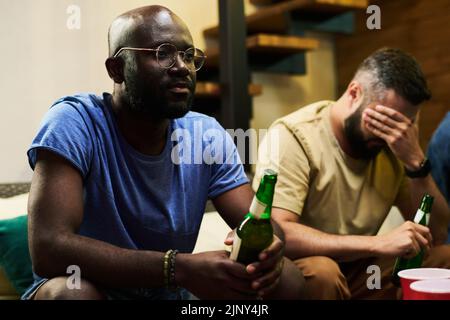 Giovane uomo afroamericano sconvolto con una bottiglia di birra guardando la trasmissione tv di partita di calcio con i suoi amici in garage Foto Stock