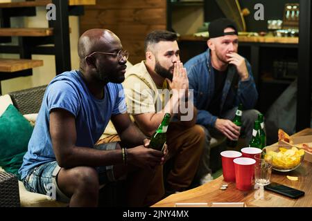 Tre giovani amici maschili interculturali che esprimono preoccupazione mentre guardano la partita di calcio trasmessa davanti al tavolo con birra e snack Foto Stock