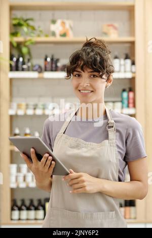 Giovane assistente femminile di successo con tablet digitale in piedi davanti alla fotocamera contro gli schermi con prodotti beautycare Foto Stock