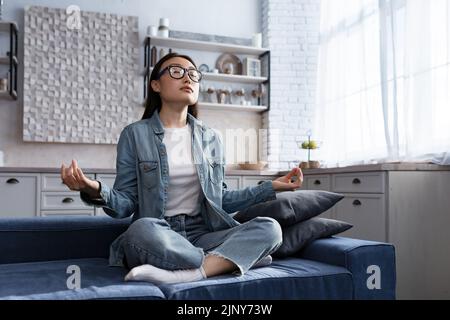 Bella donna asiatica che si rilassa a casa seduta sul divano e meditando in posizione lotus, facendo esercizi di respirazione nel soggiorno Foto Stock