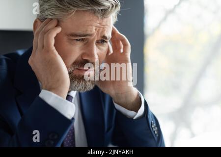 Sconvolto uomo d'affari maturo che soffre acute mal di testa sul posto di lavoro in ufficio Foto Stock