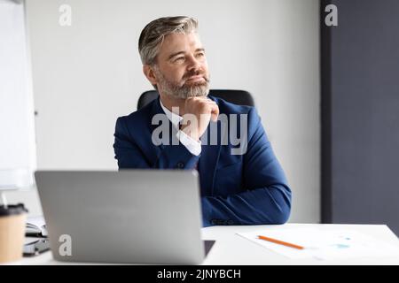 Ritratto di Dreamy uomo d'affari maturo seduto alla scrivania con laptop in ufficio Foto Stock