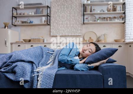 Giovane bella donna asiatica che riposa e dorme a casa sdraiata sul divano sotto coperta nel soggiorno, sonnellino Foto Stock