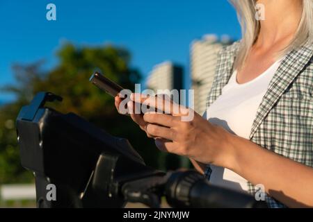 Primo piano delle mani di una donna con un telefono cellulare utilizzando un'applicazione per sbloccare uno scooter elettrico in una giornata di sole sulla strada Foto Stock