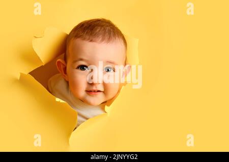 Un bambino felice guarda fuori da un buco nello studio sfondo giallo. Sorridendo il bambino sbirciando attraverso uno sfondo di carta strappato, spazio di copia. Età dei bambini un anno Foto Stock