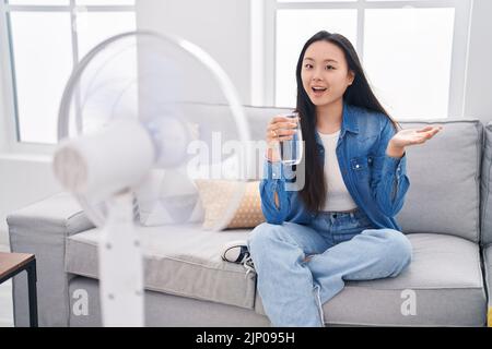 Giovane donna asiatica bere un bicchiere d'acqua godendo l'aria dal ventilatore celebrare il successo con sorriso felice e l'espressione del vincitore con mano sollevata Foto Stock