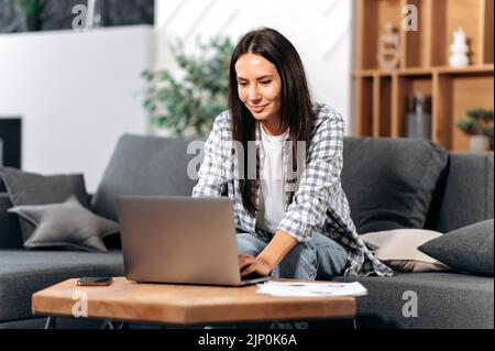 Soddisfatto successo castana caucasica donna, sedersi in salotto, utilizzando il computer portatile, lavorando su un importante progetto da casa, ha concentrato la digitazione di testo sulla tastiera, alla ricerca di informazioni su Internet Foto Stock
