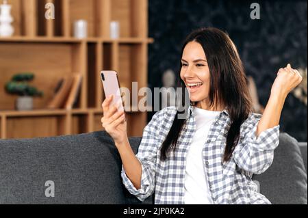 Trionfo, vinci. Allegro felice donna giovane brunetta caucasica in abbigliamento casual, sedersi a casa in soggiorno su comodo divano, utilizzando smartphone, leggere le buone notizie, gesturing con la mano, sorridendo gioiosamente Foto Stock