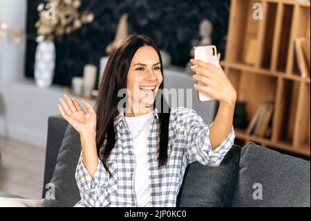 Felice ragazza caucasica in abbigliamento casual, prende un selfie al telefono. Ragazza gioiosa seduta sul divano del soggiorno, utilizza lo smartphone per comunicare tramite videoconferenza con gli amici, salutare, sorridere Foto Stock