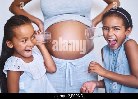 Lo sento muoversi. Ritratto di due ragazze allegre in piedi accanto alla loro madre, mentre ciascuno mettere un bicchiere sul suo ventre incinta per ascoltare Foto Stock