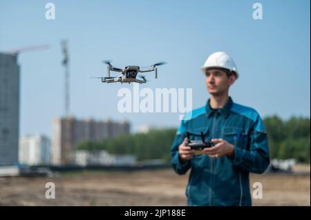 Un uomo in un casco e tute controlla un drone in un cantiere. Il costruttore esegue la supervisione tecnica. Foto Stock