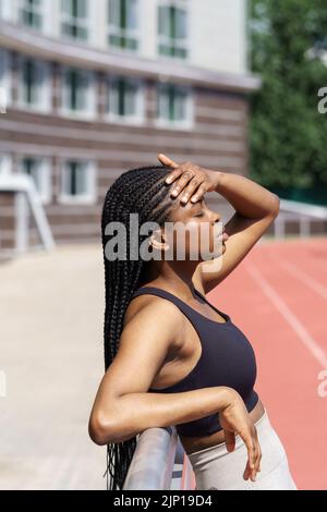 La stanca donna afroamericana riposa dopo un allenamento intensivo appoggiandosi alla recinzione metallica del parco giochi Foto Stock