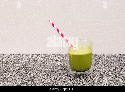 Il tè Matcha in una tazza di vetro con una paglia rossa si erge su un acciaio e sullo sfondo un fondo sfocato della cucina Foto Stock