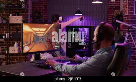 Giocatore che esegue lo sparatutto in prima persona in streaming sul pc, mentre la donna utilizza occhiali vr per la simulazione in salotto. Uomo che gioca a gioco d'azione online multiplayer mentre la ragazza di gioco sta combattendo in gioco di realtà virtuale. Foto Stock