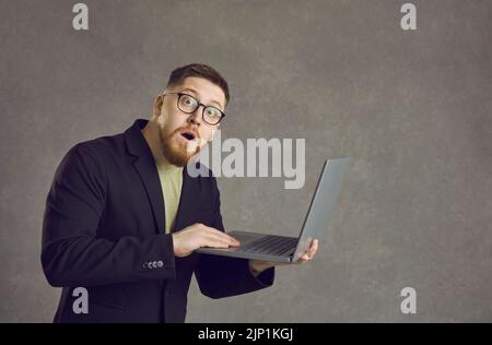 Ritratto di un barbuto scioccato uomo in piedi con un computer portatile in mano su uno sfondo grigio. Foto Stock