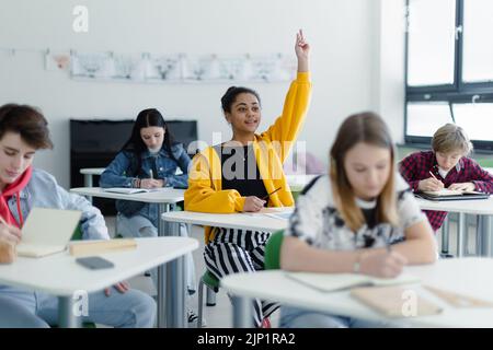 Gli studenti delle scuole superiori prestano attenzione in classe, seduti sulle loro scrivanie e alzando la mano. Foto Stock