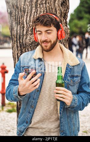 Giovane hipster che ascolta la musica con le cuffie wireless all'aperto o guardare musica media video con il suo smartphone cellulare bere una birra o Foto Stock